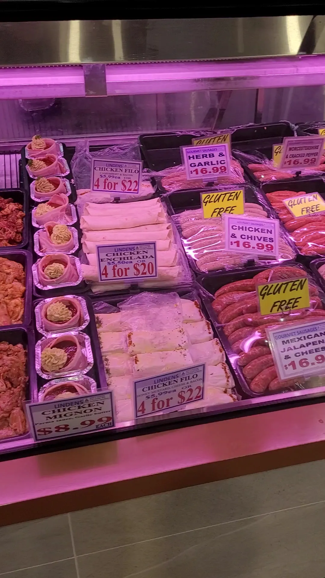 Refrigerated counter display showing chicken filo rolls, chicken enchiladas, gourmet sausages with handwritten price signs under pink lighting