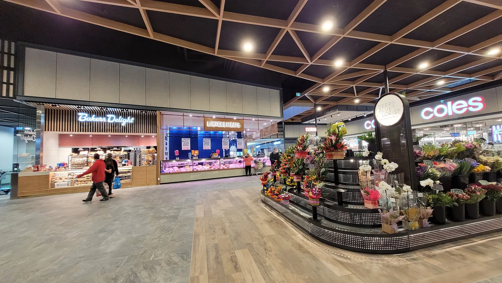 Wide-angle view of the Karingal Hub fresh food precinct with Lindens Meats visible in the background alongside a florist and adjacent food stalls, shoppers walking through the corridor